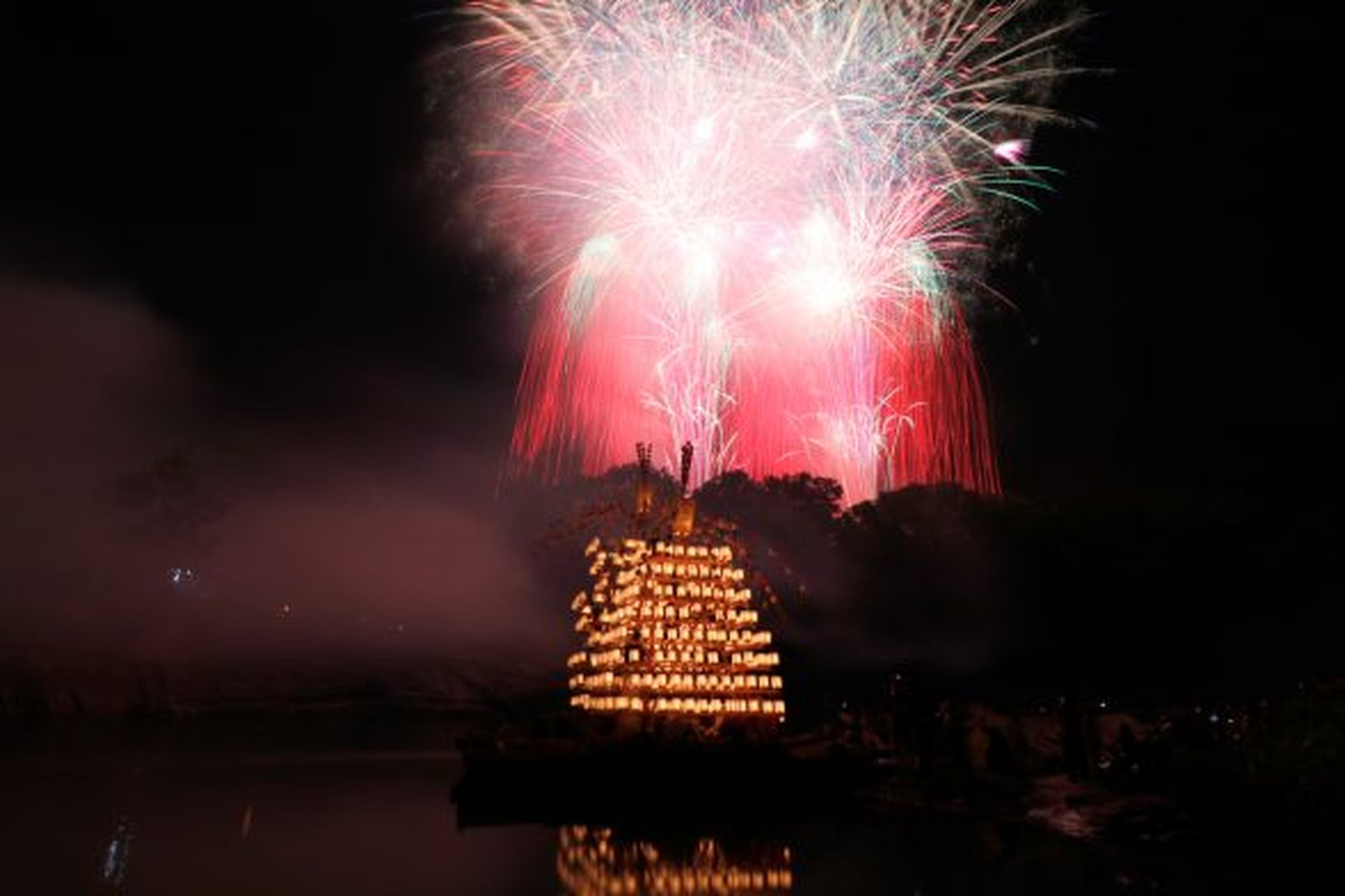 長瀞船玉まつりで、赤や緑の花火が打ち上がる中、灯りをともした船が水面に映る様子