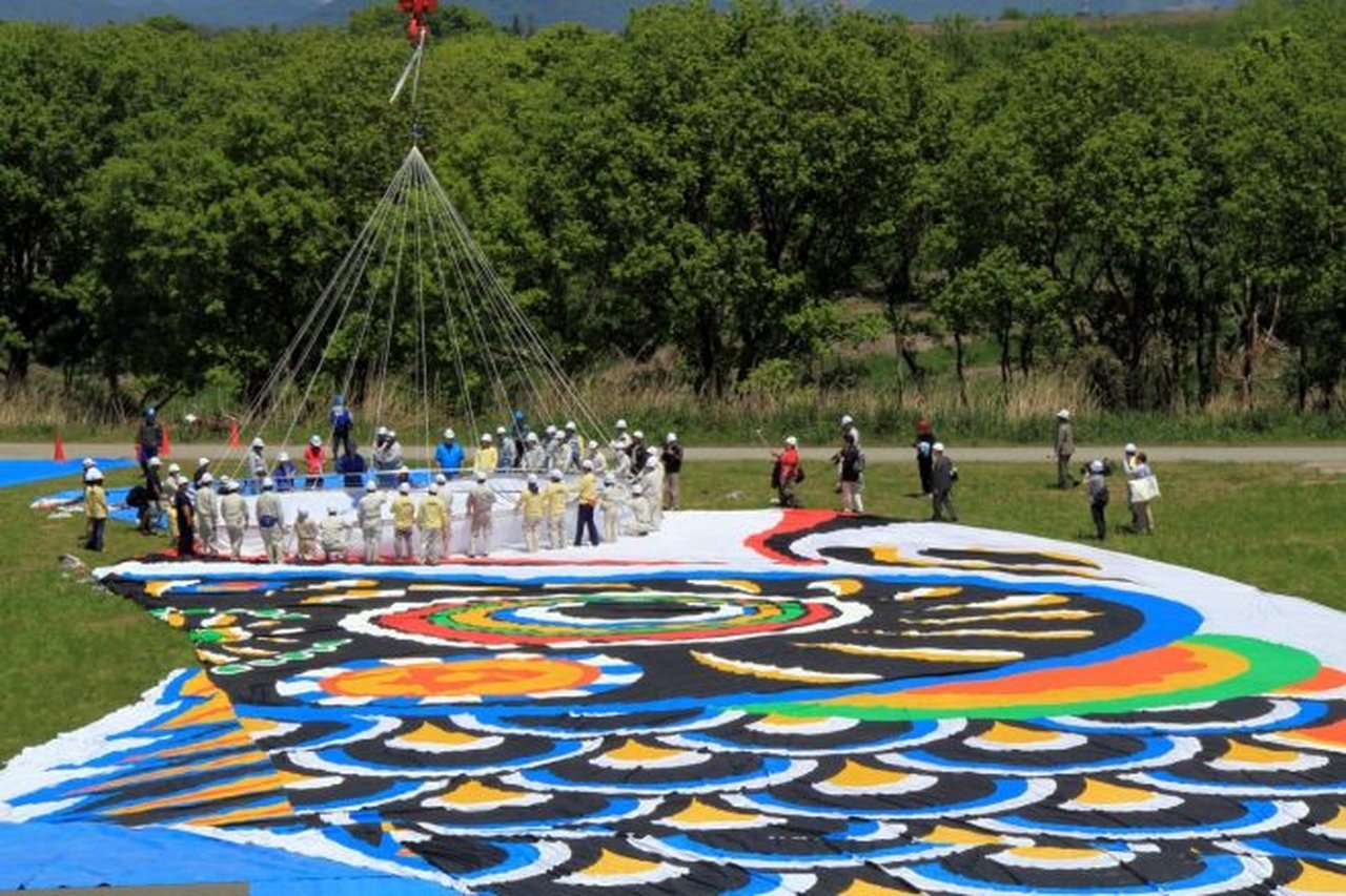 地面いっぱいに広げられたジャンボこいのぼりの布を、スタッフが多数で整える壮大な準備風景