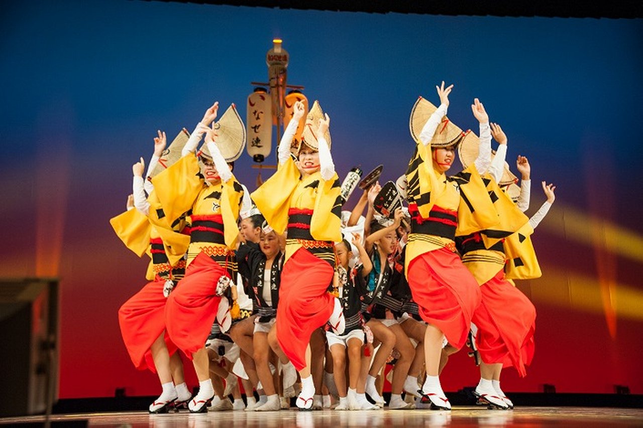 南越谷阿波踊りの舞台で、黄色と赤の衣装の子ども連が夕景ライトの中で力強く踊る場面