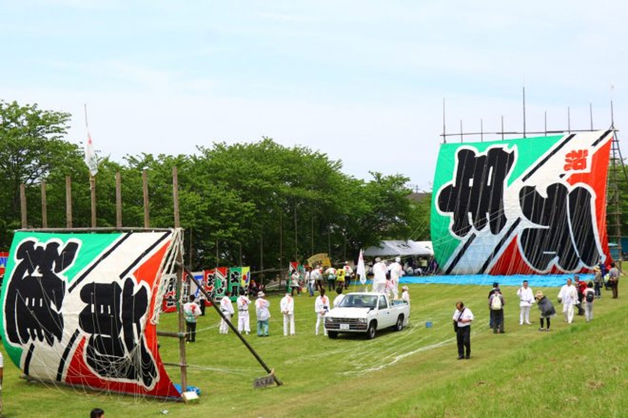 揚げ場に立てられた複数の巨大凧と、祭り会場に集まる参加者