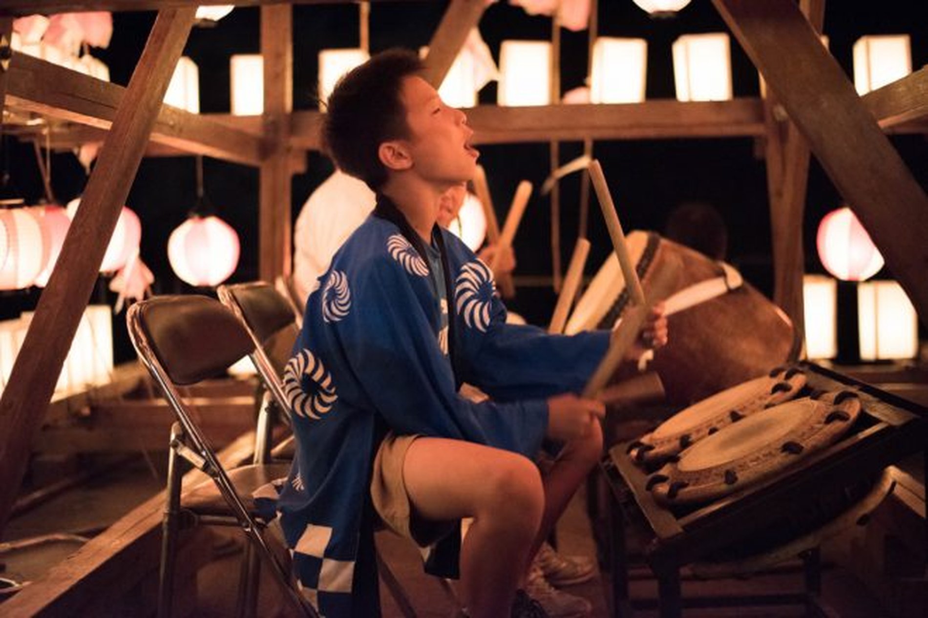 提灯に照らされながら、万灯の上で太鼓を力強く叩く少年の姿